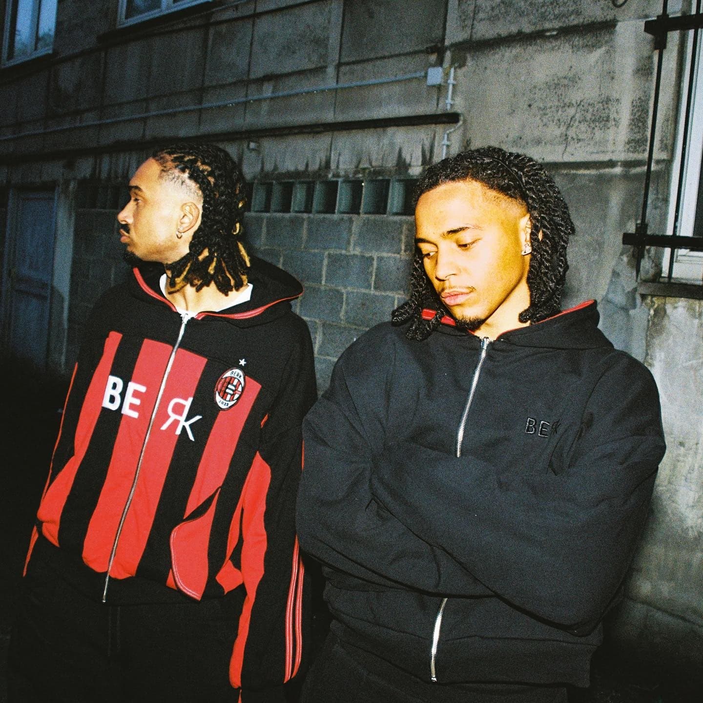 BERK STUDIOS campaign: two models against a concrete wall, one in a red and black striped zip jacket carrying the BERK wordmark and crest, one in a black hooded jacket with an embroidered BERK mark.